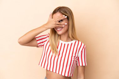 Sarışın Uruguaylı kız bej arka planda izole edilmiş gözleri elleriyle kaplayan ve gülümseyen.