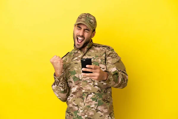 Military man isolated on yellow background with phone in victory position