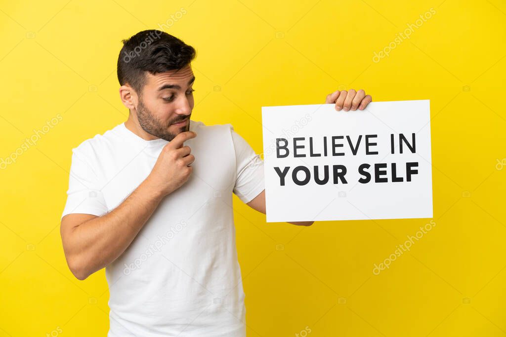 Young handsome caucasian man isolated on yellow background holding a placard with text Believe In Your Self and thinking