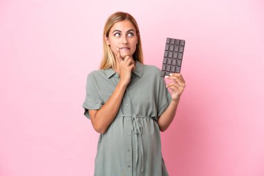 Genç Uruguaylı kadın mavi arka planda izole edilmiş hamile ve elinde çikolata tutarken şüpheleri var.