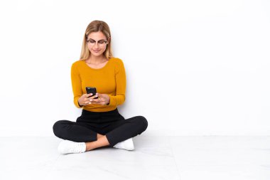 Sarışın Uruguaylı kız yerde oturuyor ve cep telefonuyla mesaj yolluyor.