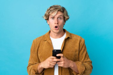 İngiliz adam mavi arka plan üzerinde izole edilmiş kameraya bakarken şaşırmış bir ifadeyle cep telefonunu kullanıyor.