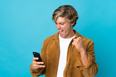 İngiliz adam mavi arka planda cep telefonu kullanıyor ve zafer jesti yapıyor.