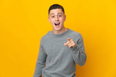 Sarı arka planda izole edilmiş genç beyaz adam şaşırmış ve ön tarafı işaret ediyor.