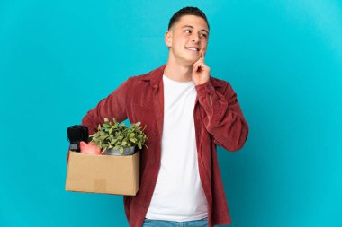 Kafkas 'lı genç bir adam mavi arka planda izole edilmiş şeylerle dolu bir kutuyu kaldırırken bir yandan yukarı bakarken bir fikir düşünüyor.