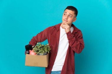 Kafkasyalı genç bir adam mavi arka planda izole edilmiş eşyalarla dolu bir kutuyu alıp...