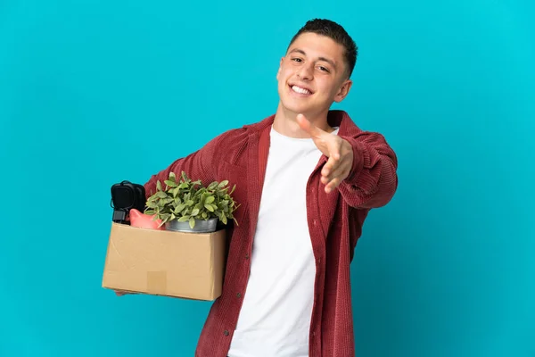 Young caucasian man making a move while picking up a box full of things isolated on blue background shaking hands for closing a good deal