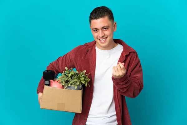Young caucasian man making a move while picking up a box full of things isolated on blue background making money gesture