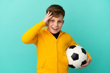 Mavi arka planda futbol oynayan kızıl saçlı çocuk sürpriz bir ifadeyle izole edilmiş.