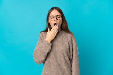 Genç Litvanyalı bir kadın mavi arka planda izole edilmiş esniyor ve ağzını eliyle açık tutuyor.