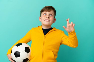 Kızıl saçlı çocuk mavi arka planda izole bir şekilde futbol oynuyor. Şans diliyor ve en iyisini diliyor.