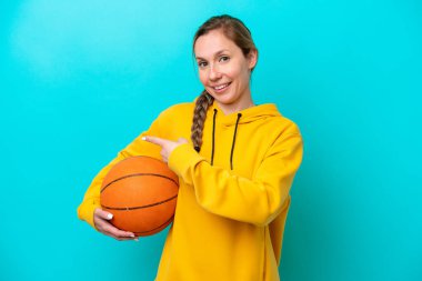 Genç beyaz kadın mavi arka planda izole edilmiş basketbol oynuyor ve yan tarafı işaret ediyor.