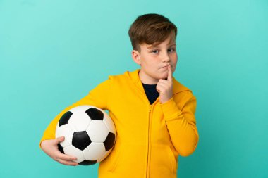 Kızıl saçlı çocuk mavi arka planda izole bir şekilde futbol oynuyor.