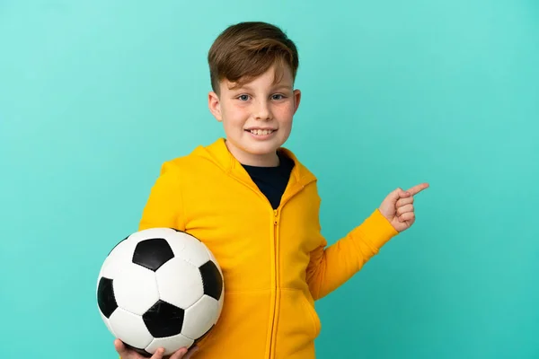 Redhead Kid Playing Football Isolated Blue Background Pointing Finger Side — Stockfoto