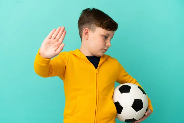 Redhead kid playing football isolated on blue background making stop gesture and disappointed
