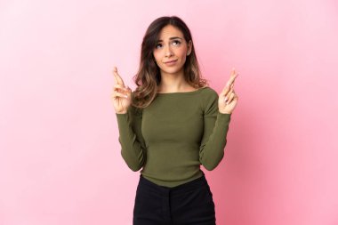 Pembe arka planda izole edilmiş genç beyaz kadın parmaklarını çapraz yapıp en iyisini diliyor.