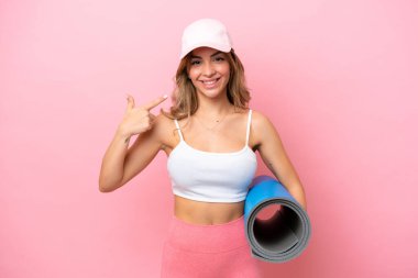 Genç sporcu kadın, elinde minderle yoga derslerine gidiyor ve baş parmak hareketi yapıyor.