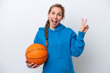 Beyaz arka planda izole edilmiş basketbol oynayan beyaz kadın gülümsüyor ve zafer işareti gösteriyor.