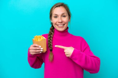 Elinde kızarmış cips tutan genç beyaz kadın mavi arka planda izole edilmiş bir ürün sunmak için yan tarafı işaret ediyor.
