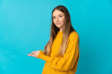 Genç Litvanyalı bir kadın mavi arka planda izole edilmiş. Davet etmek için ellerini yana uzatıyor.