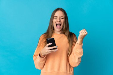 Genç Litvanyalı kadın mavi arka planda cep telefonu kullanıyor ve zafer jesti yapıyor.