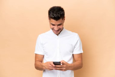 Beyazlı genç adam bej arka planda izole edilmiş. Cep telefonuyla mesaj gönderiyor.