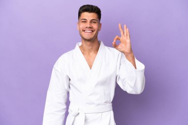 Mor arka planda karate yapan genç beyaz adam parmaklarıyla tamam işareti gösteriyor.