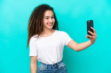 Mavi arka planda izole edilmiş genç bir Arap kadın cep telefonuyla selfie çekiyor.