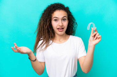 Görünmez diş telleri tutan genç bir Arap kadın mavi arka planda şok olmuş yüz ifadesiyle izole edilmiş.