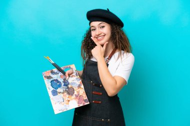 Genç sanatçı Arap kadın mavi arka planda izole edilmiş bir palet tutuyor mutlu ve gülümsüyor.