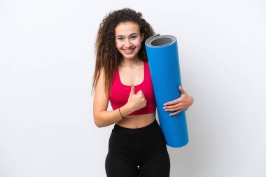 Genç Arap spor kadını yoga derslerine giderken beyaz arka planda izole edilmiş bir minder tutuyor ve baş parmak hareketi yapıyor.
