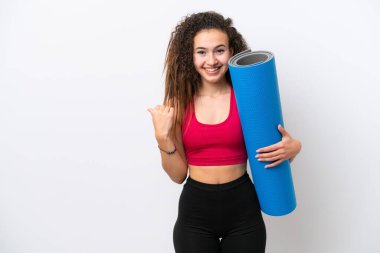 Genç Arap spor kadını yoga derslerine giderken beyaz arka planda izole edilmiş bir minder tutuyor ve bir ürünü sunmak için yan tarafı işaret ediyor.