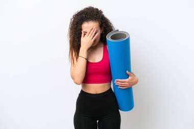 Genç Arap spor kadını yoga derslerine giderken beyaz bir zeminde yorgun ve hasta bir ifadeyle izole edilmiş bir minder tutuyor.