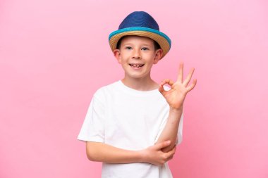 Pembe arka planda izole edilmiş şapka takan küçük beyaz çocuk parmaklarıyla tamam işareti gösteriyor.
