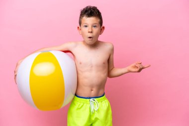 Küçük beyaz çocuk elinde bir plaj topu tutuyor. Pembe arka planda izole edilmiş. Şaşırmış ve işaret ediyor.