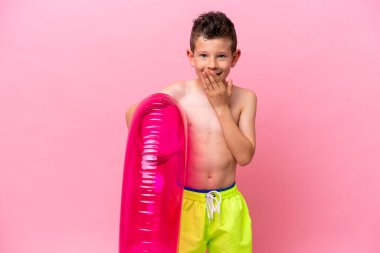 Küçük beyaz çocuk pembe arka planda izole edilmiş şişirilebilir bir çörek tutuyor mutlu ve ağzı eliyle kaplayan bir gülümseme.