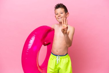 Küçük beyaz çocuk pembe arka planda izole edilmiş şişme bir çörek tutuyor ve parmaklarıyla üçe kadar sayıyor.