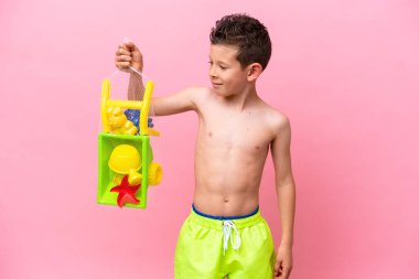 Küçük beyaz çocuk elinde kumsal oyuncakları tutarken mutlu bir ifadeyle pembe arka planda izole edilmiş.