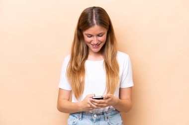 Bej arka planda izole edilmiş genç beyaz kadın cep telefonuyla bir mesaj gönderiyor.