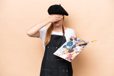 Beyaz tenli genç bir kadın bej arka planda izole edilmiş bir palet tutuyor gözlerini elleriyle kapıyor. Bir şey görmek istemiyorum.