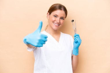 Beyaz dişçi kadının elinde bej arka planda izole edilmiş aletler var. Başparmakları havada çünkü iyi bir şey oldu.