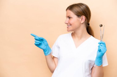 Beyaz dişçi kadının elinde bej arka planda izole edilmiş aletler var. Parmakla işaret ediyor ve bir ürün sunuyor.
