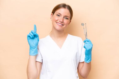 Beyaz dişçi kadının elinde bej arka planda izole edilmiş aletler var ve en iyinin işareti olarak parmağını kaldırıyor.