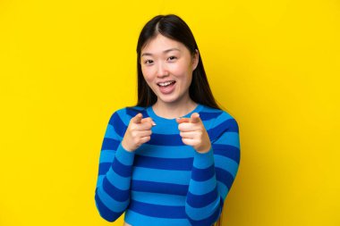 Sarı arka planda izole edilmiş genç Çinli kadın şaşırmış ve ön tarafı işaret ediyor.