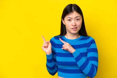 Sarı arka planda izole edilmiş genç Çinli kadın korkmuş ve kenarı işaret ediyor.