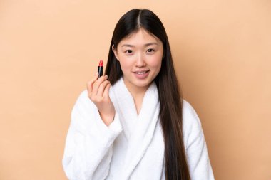 Young Chinese woman isolated on beige background holding red lipstick