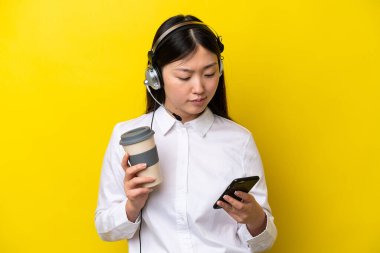 Tele pazarlamacı Çinli bir kadın sarı arka planda izole edilmiş bir kulaklıkla çalışıyor. Elinde kahve ve cep telefonu var.