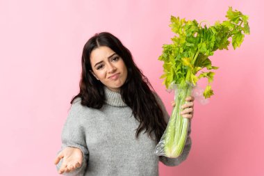 Mavi arka planda izole edilmiş bir kereviz tutan genç beyaz kadın omuzlarını kaldırırken şüpheyle hareket ediyor.