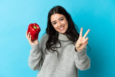 Mavi arka planda biber tutan genç beyaz kadın gülümsüyor ve zafer işareti gösteriyor.