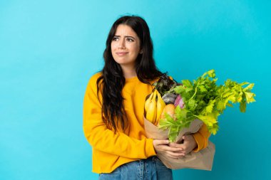 Elinde alışveriş çantası tutan genç bir kadın mavi arka planda izole edilmiş şüpheleri ve düşünceleri var.